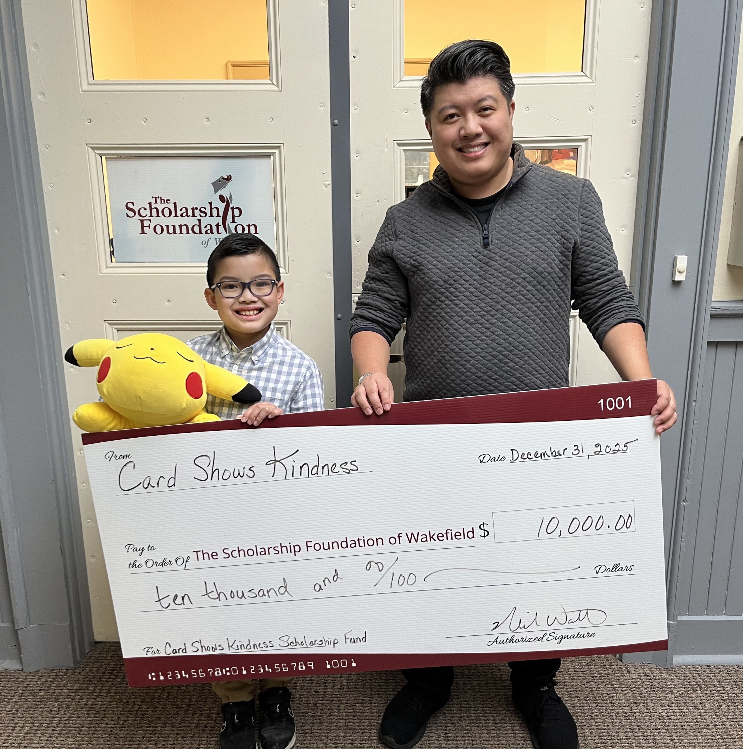 A child holding a Pikachu plush and an adult stand outside The Scholarship Foundation of Wakefield, presenting a large $10,000 donation check from Card Shows Kindness.