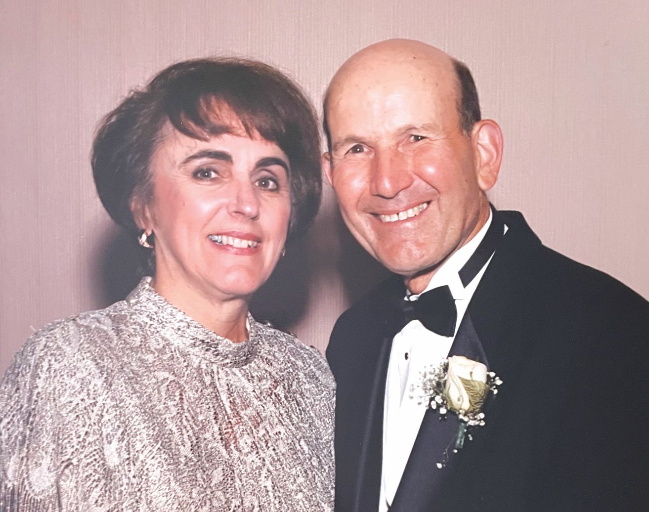 A smiling couple dressed in formal evening attire, standing close together and facing the camera. The woman is wearing a light-colored, textured jacket and earrings. The man is wearing a black tuxedo with a bow tie and a white rose boutonniere